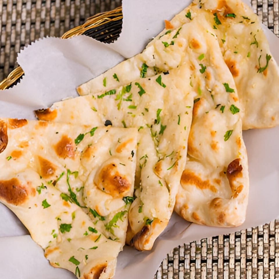 Golden-brown naan bread, garnished with fresh green herbs, served in a basket lined with white paper.