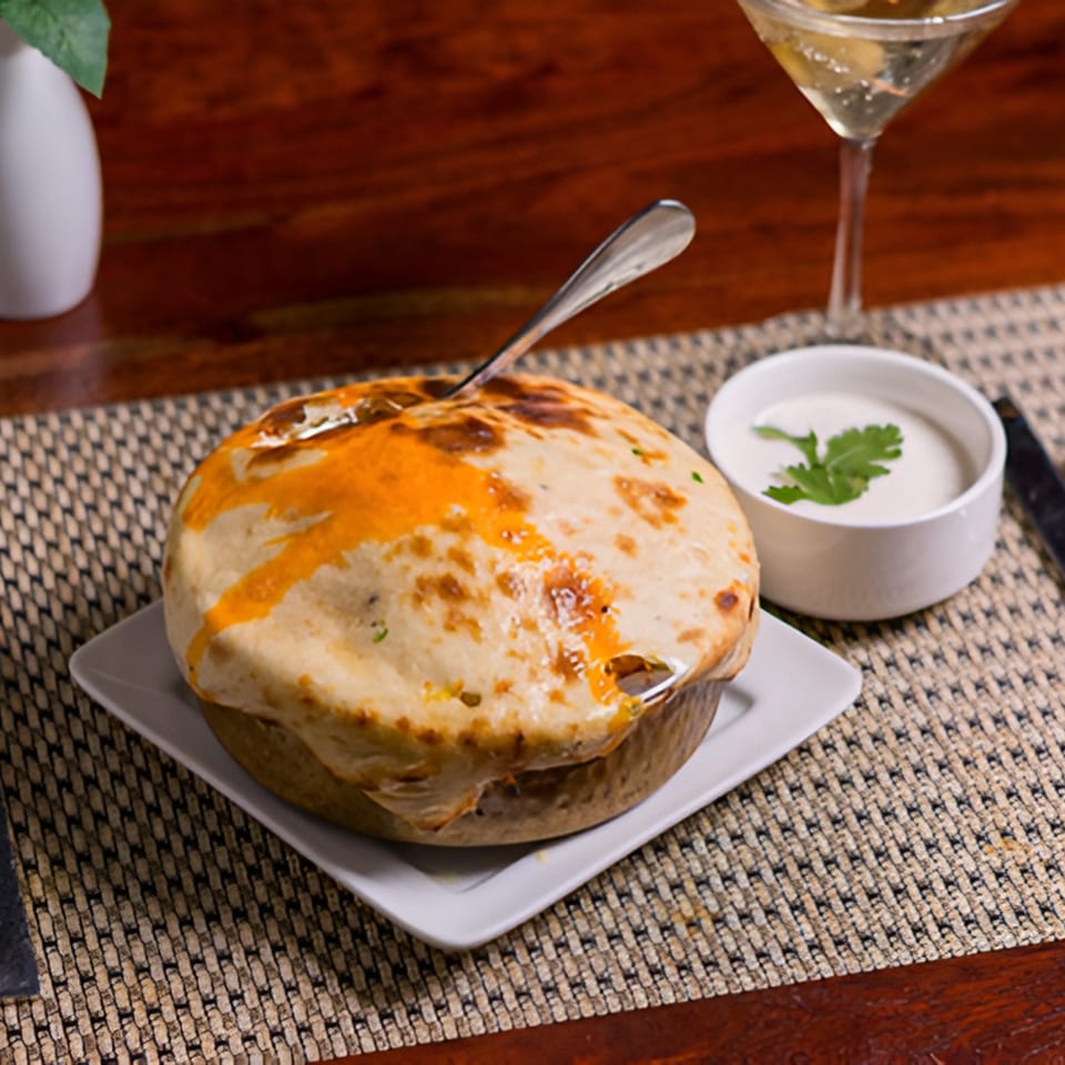Awadhi biryani with a pastry crust, served with raita and a martini on a wooden table.