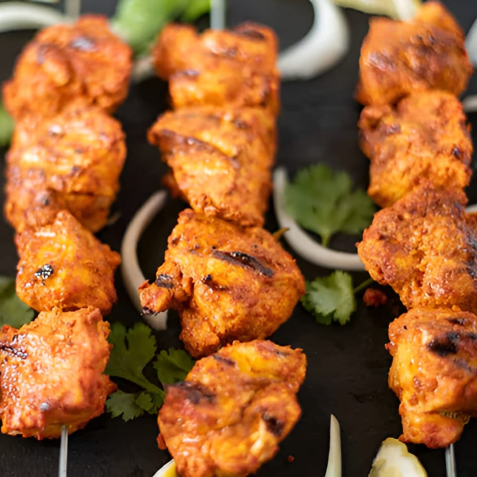 Three skewers of vibrant orange chicken tikka, garnished with fresh cilantro and onion slices.