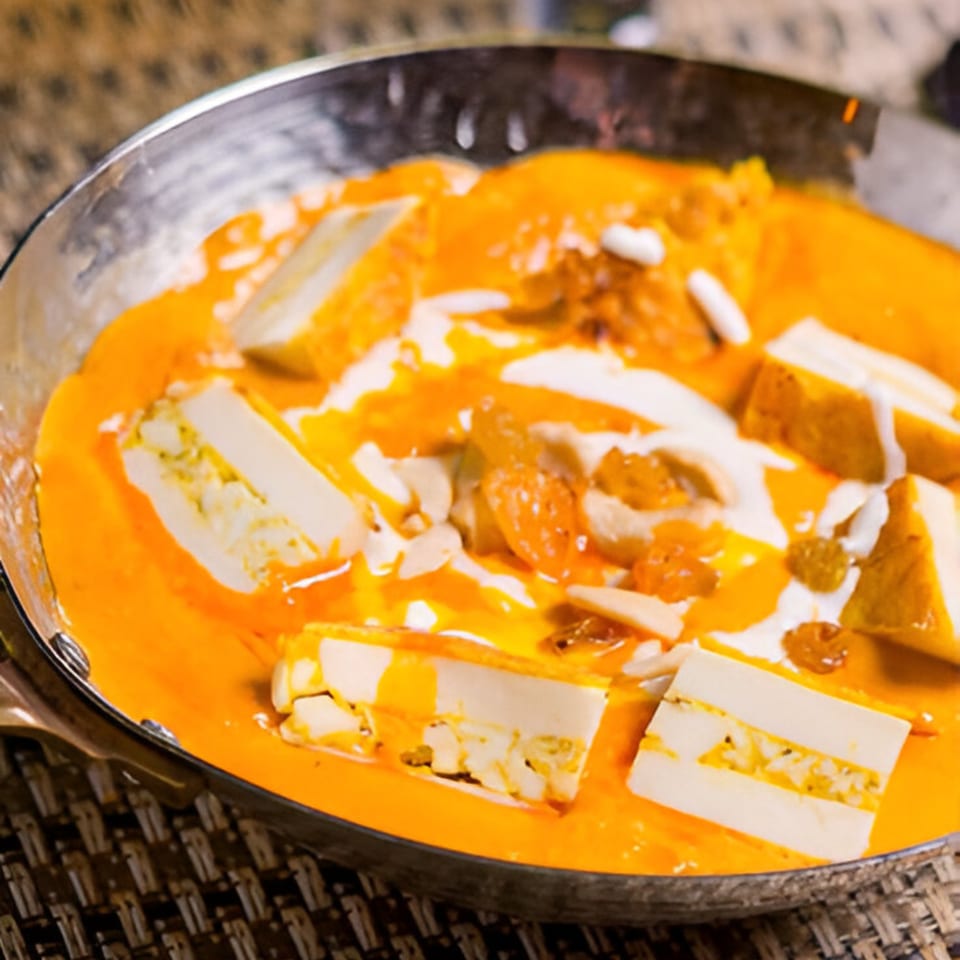 Indian paneer dish in a metal bowl, with orange gravy, white cream, raisins, and nuts.