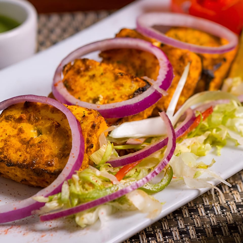 Indian paneer tikka, garnished with red onion rings and fresh lettuce, on a white plate.