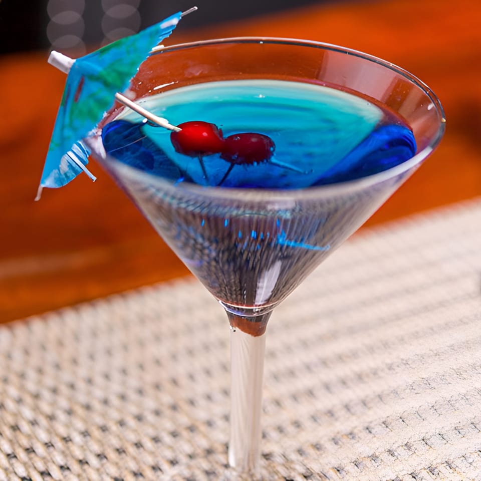 A blue cocktail in a martini glass, garnished with two cherries and a small blue umbrella.