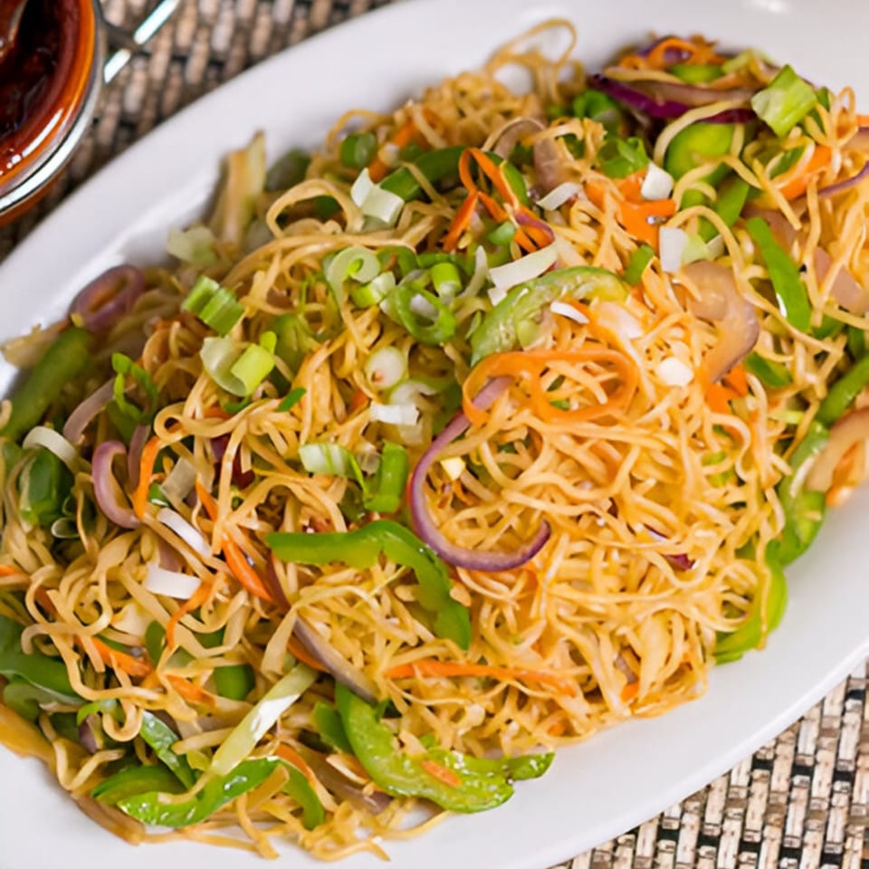 A close-up of Indian noodles with green peppers, red onions, and carrots on a white plate.